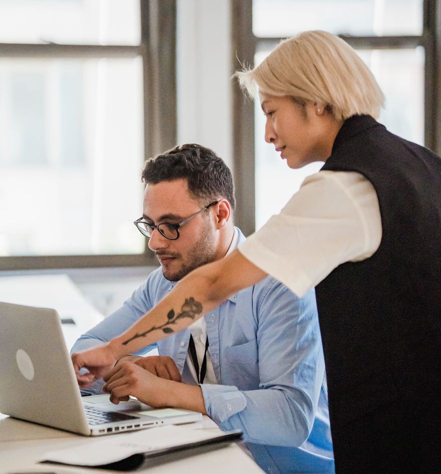A standing colleague leans in over a seated colleague's desk and reaches to his open laptop, showing him something on the screen while he follows along.